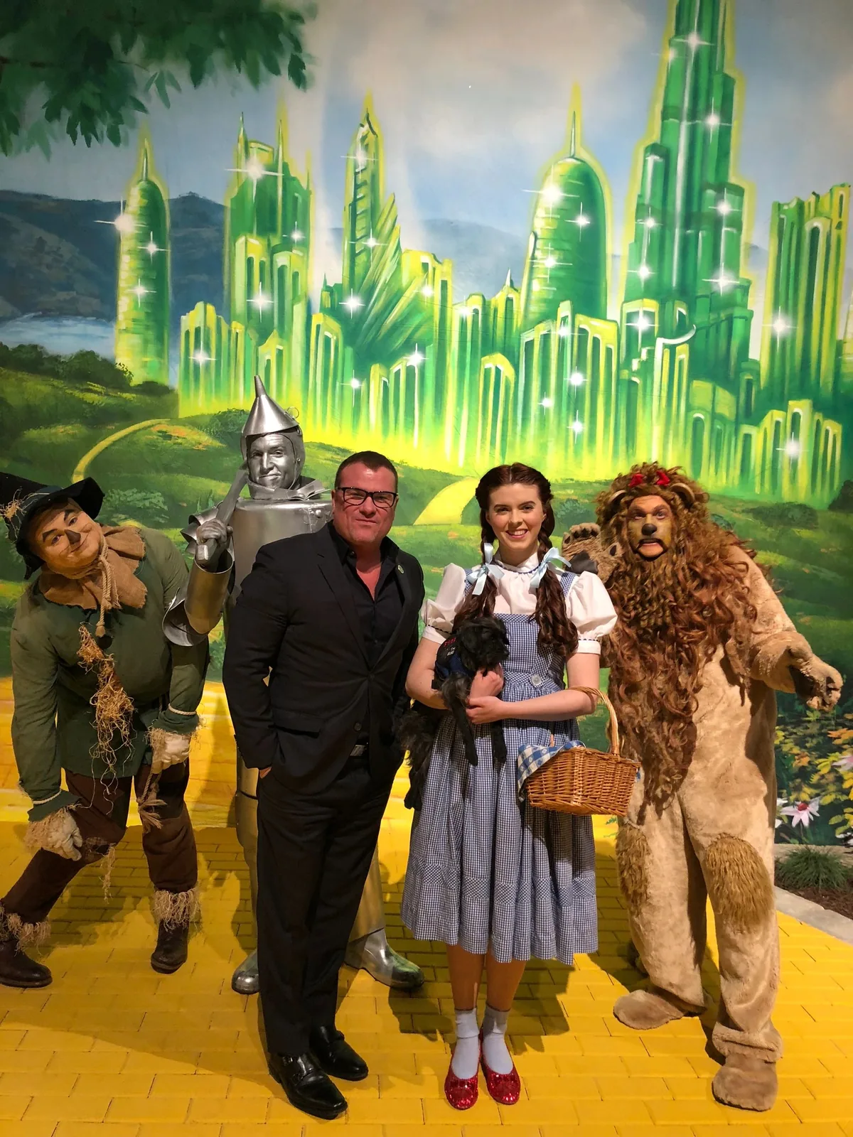 A man in black with costumed Wizard of Oz characters in front of an Emerald City mural