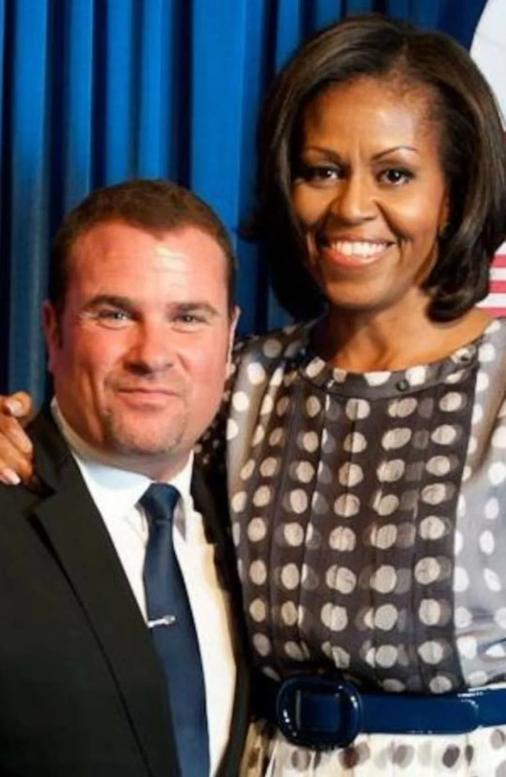 Two people posing together in front of a blue curtain and a flag