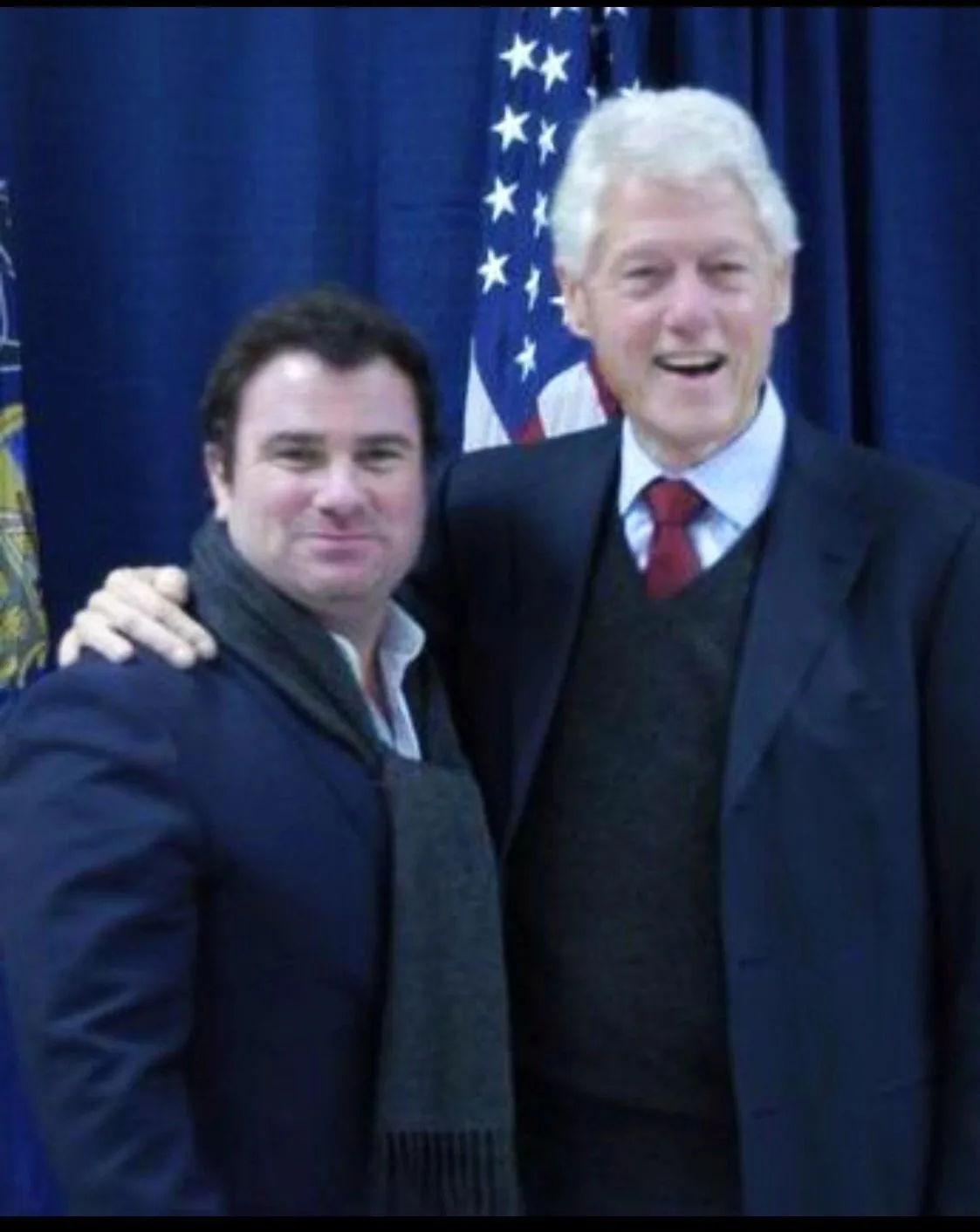 Two people posing together in front of a flag and a blue curtain